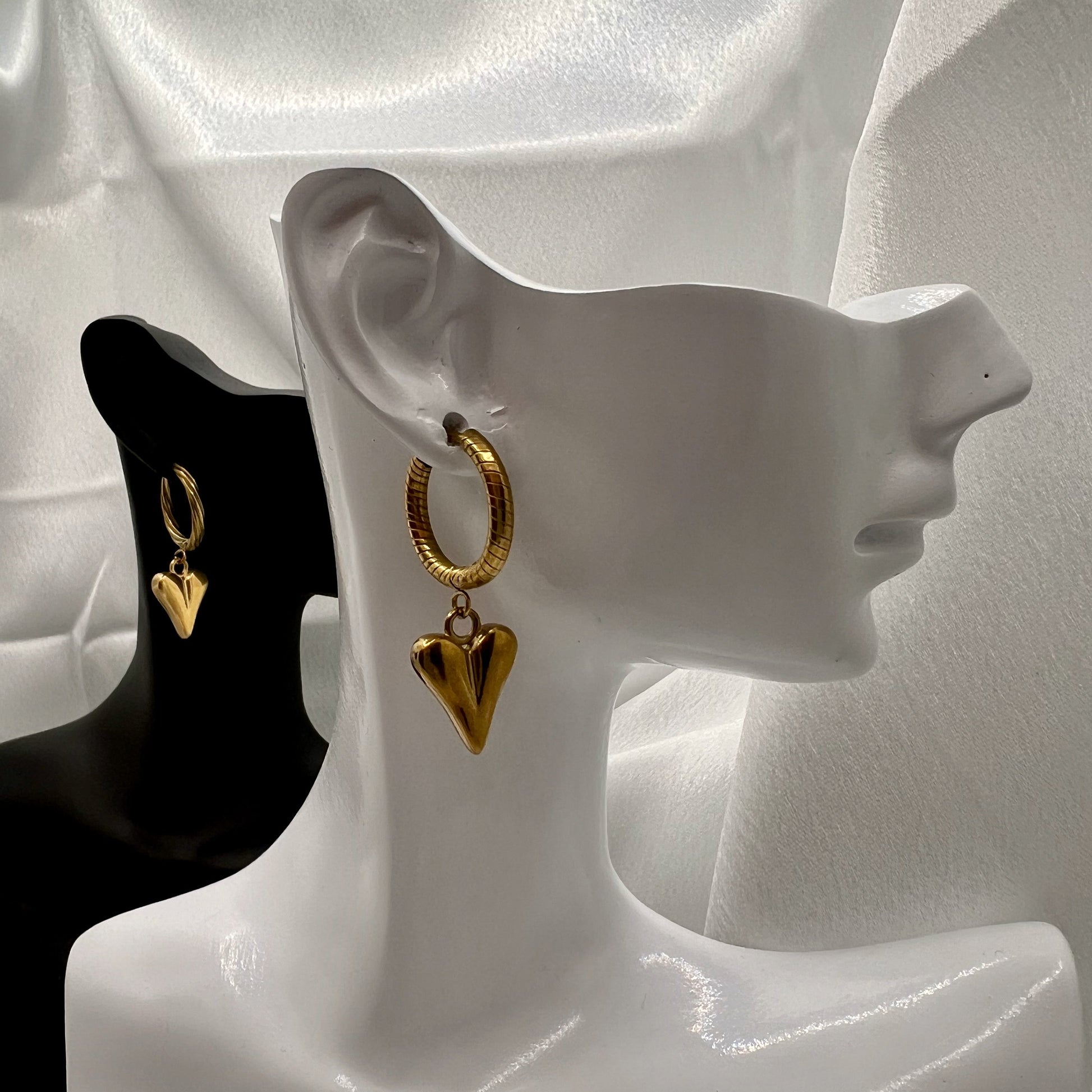 Gold heart-shaped earrings on a white mannequin ear with a black stand in the background.