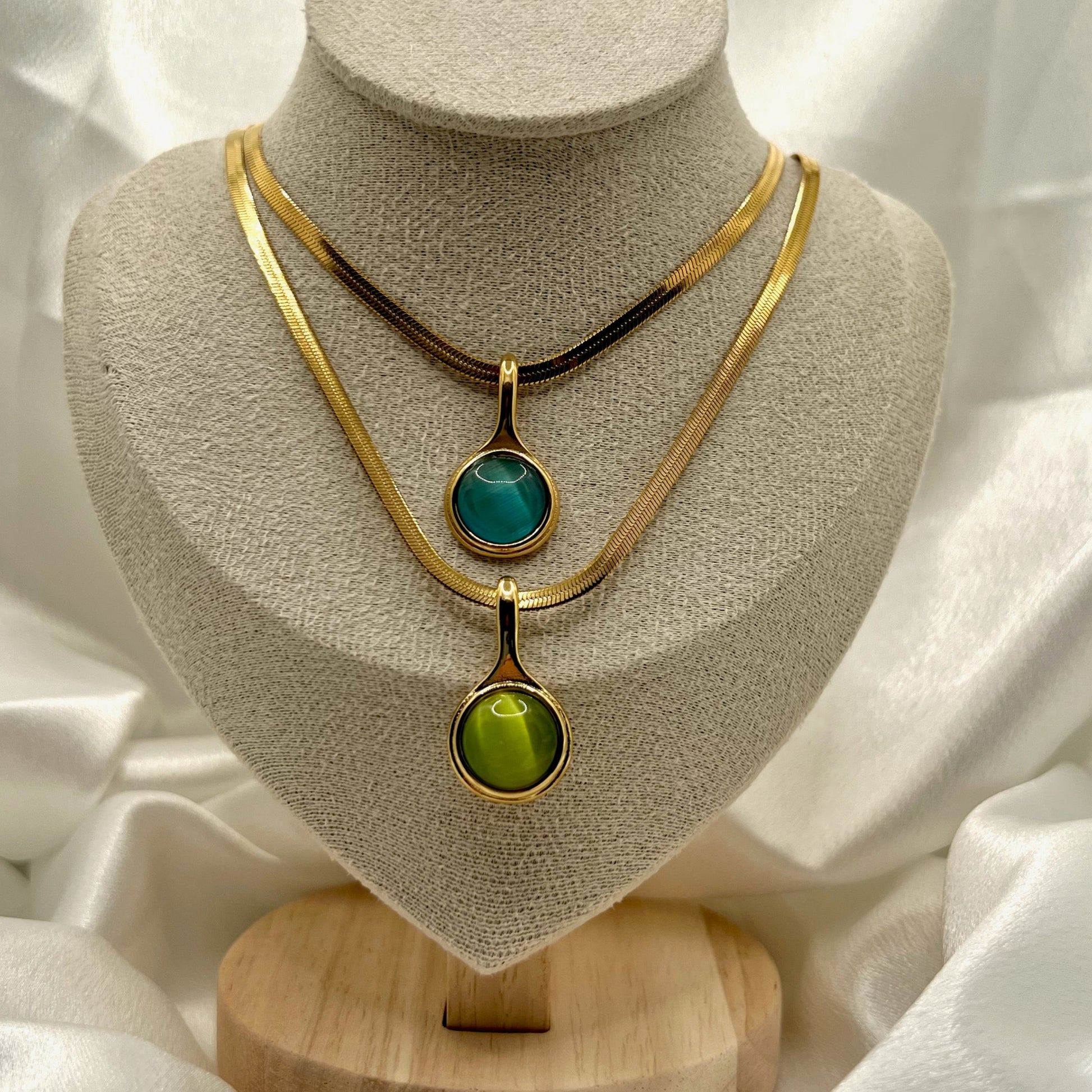 Two gold necklaces with green  and blue pendants on a beige mannequin bust against a white fabric background.