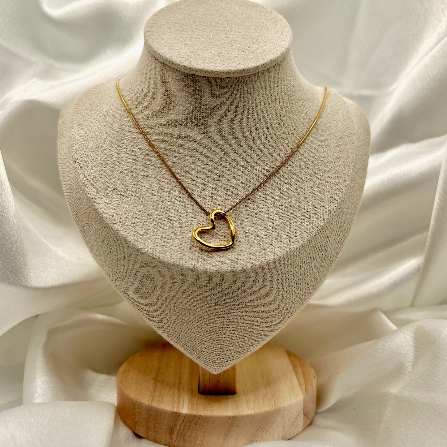 Gold heart-shaped necklace on a beige bust with a wooden base against a white fabric background