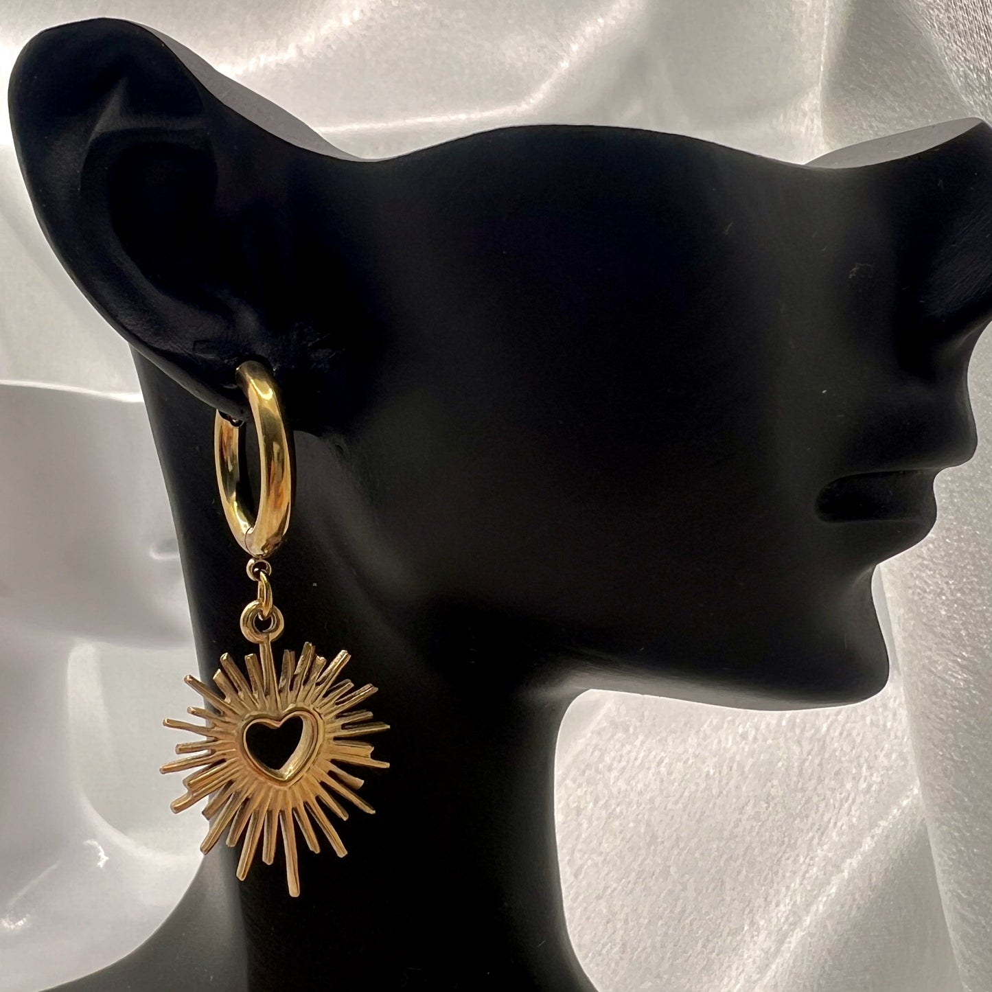 Gold earring with sunburst design on a black mannequin ear against a white background