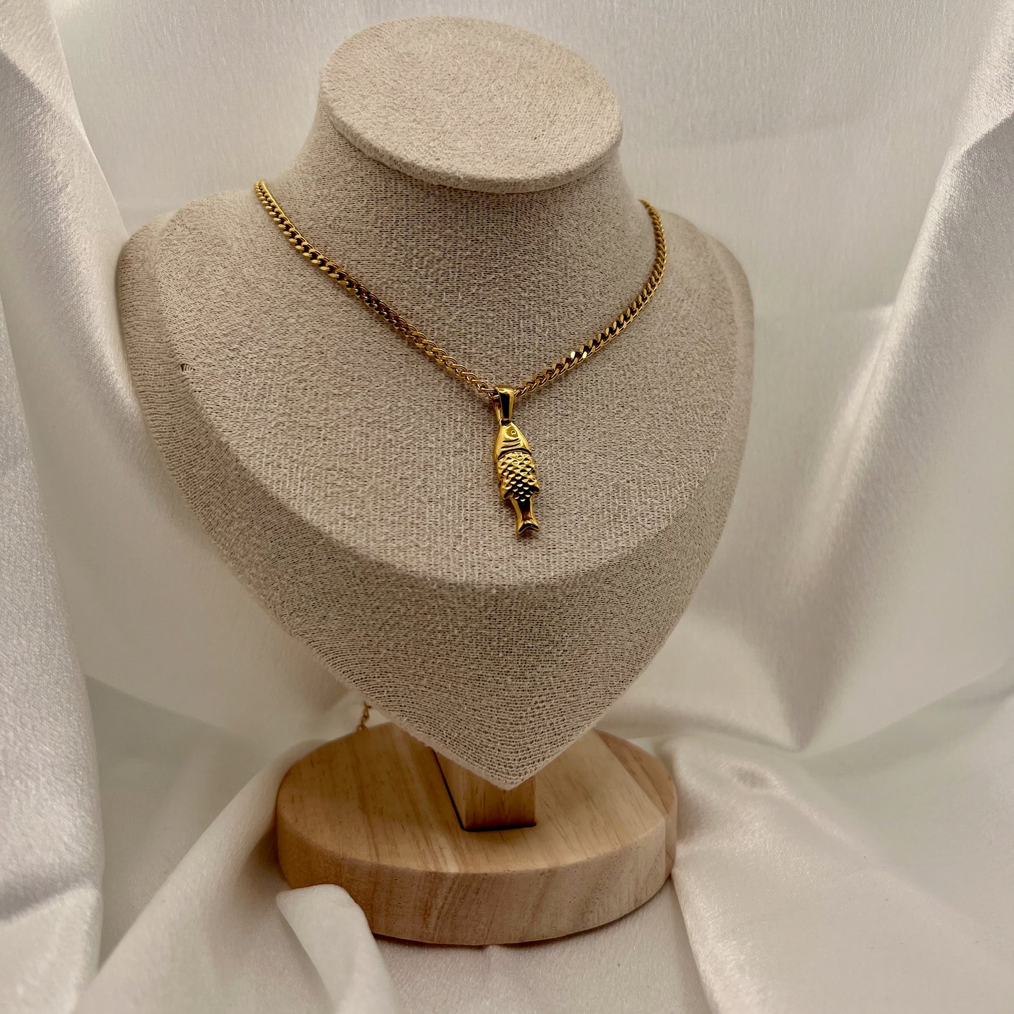 Gold necklace with a fish pendant on a beige jewelry display stand against a white fabric background